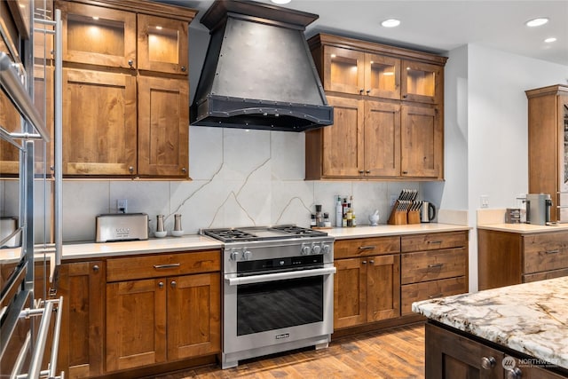 kitchen featuring light stone countertops, light wood-type flooring, premium range hood, tasteful backsplash, and stainless steel range