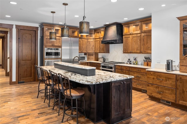 kitchen featuring dark hardwood / wood-style flooring, premium range hood, high end appliances, hanging light fixtures, and an island with sink