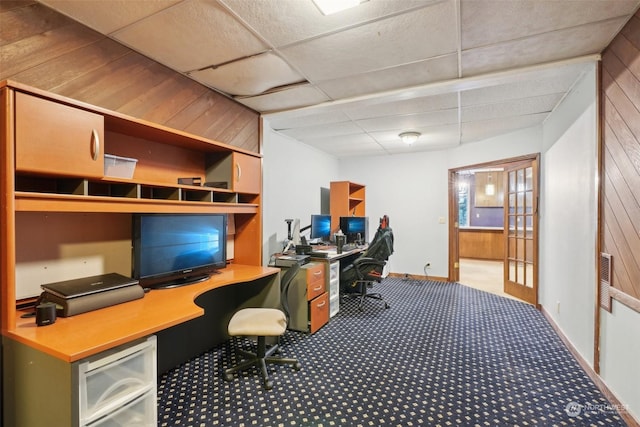 carpeted office space with french doors, built in desk, a drop ceiling, and wooden walls