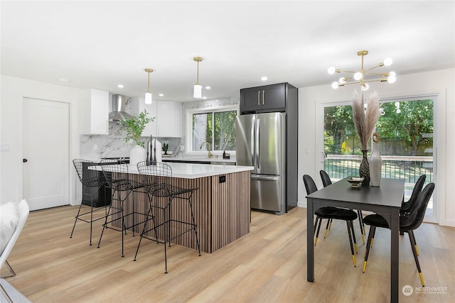 kitchen with stainless steel refrigerator, white cabinetry, wall chimney range hood, a kitchen breakfast bar, and pendant lighting