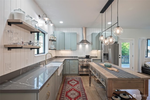 kitchen with dark stone counters, sink, hanging light fixtures, wall chimney exhaust hood, and stainless steel appliances