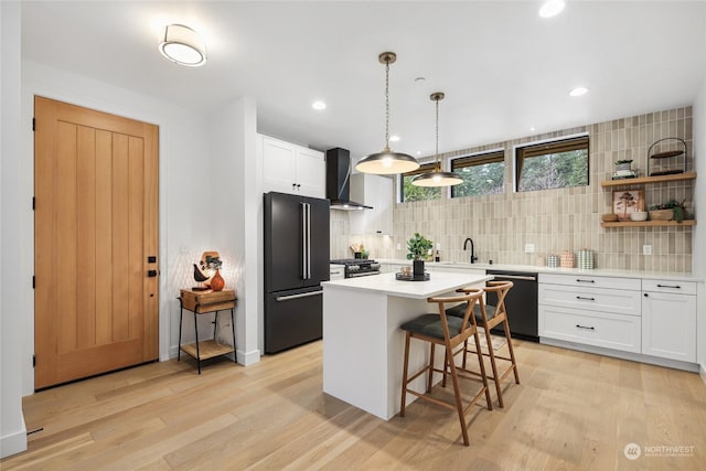 kitchen featuring hanging light fixtures, wall chimney range hood, white cabinets, a kitchen island, and appliances with stainless steel finishes