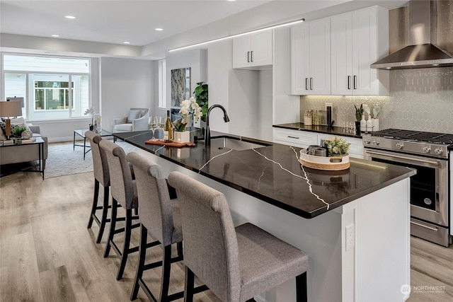 kitchen featuring wall chimney exhaust hood, stainless steel gas range, white cabinetry, a center island with sink, and a kitchen breakfast bar