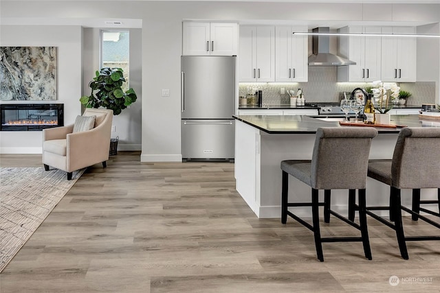 kitchen with high quality fridge, white cabinets, a kitchen breakfast bar, light wood-type flooring, and wall chimney exhaust hood