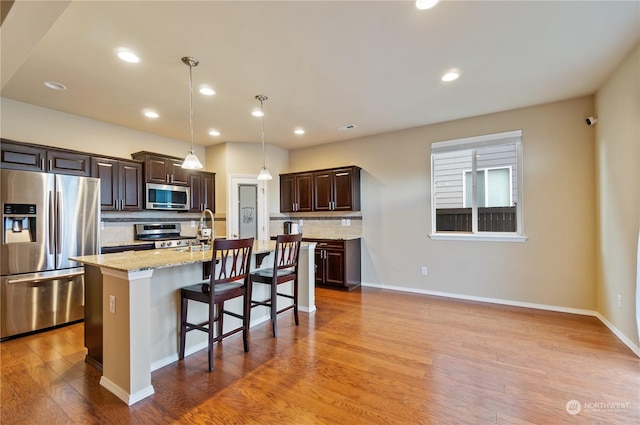 kitchen with hanging light fixtures, decorative backsplash, an island with sink, stainless steel appliances, and light stone counters