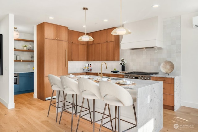 kitchen with premium range hood, decorative backsplash, an island with sink, decorative light fixtures, and light hardwood / wood-style floors