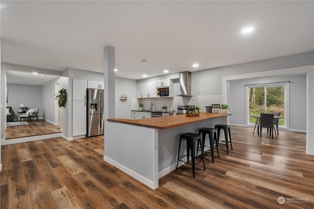 kitchen with kitchen peninsula, stainless steel appliances, wall chimney range hood, white cabinets, and butcher block countertops