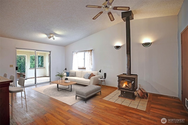 living room featuring vaulted ceiling, a wood stove, wood finished floors, a textured ceiling, and ceiling fan