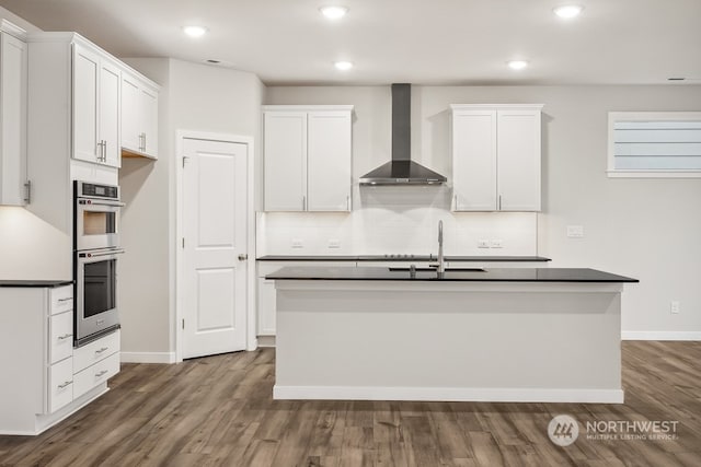 kitchen featuring white cabinetry, double oven, wall chimney exhaust hood, and a center island with sink