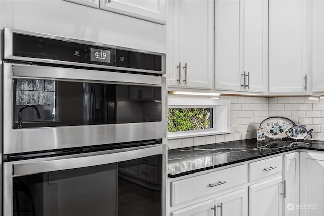 kitchen featuring stainless steel double oven, backsplash, dark stone countertops, and white cabinets