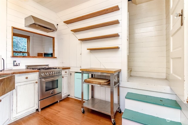 kitchen featuring wood walls, wall chimney range hood, stainless steel range, light wood-type flooring, and white cabinets