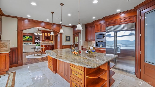 kitchen featuring crown molding, hanging light fixtures, built in appliances, ornate columns, and an island with sink