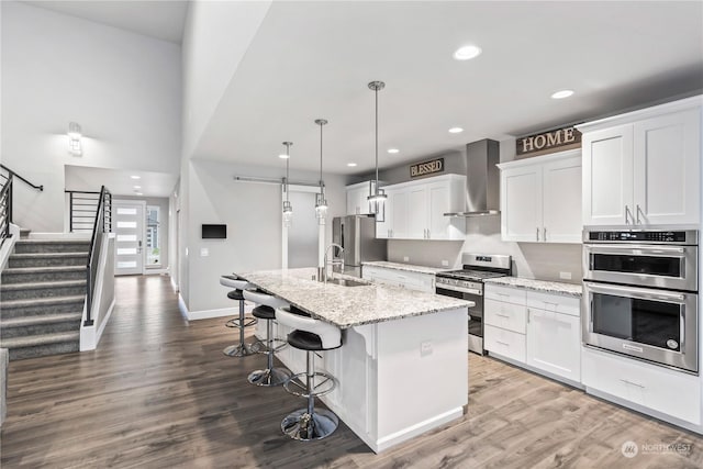 kitchen with light stone counters, wall chimney range hood, stainless steel appliances, a kitchen island with sink, and white cabinets