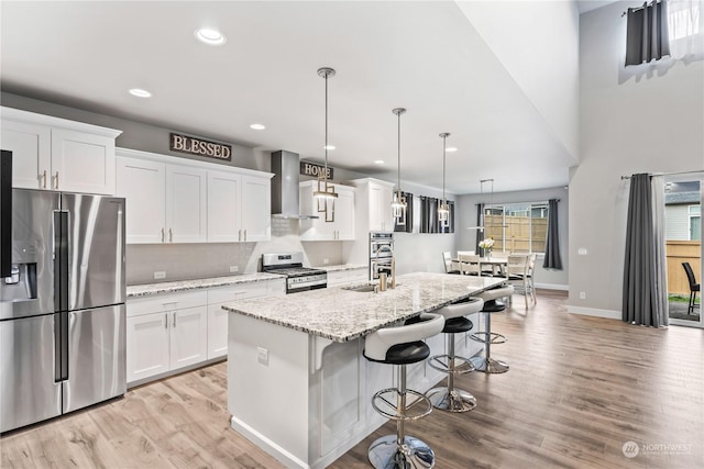 kitchen with decorative light fixtures, white cabinets, a kitchen island with sink, stainless steel appliances, and wall chimney range hood