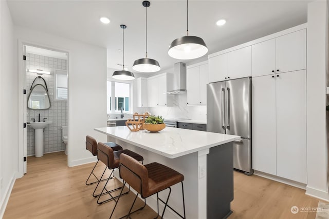 kitchen with high end refrigerator, wall chimney exhaust hood, pendant lighting, white cabinets, and a kitchen island