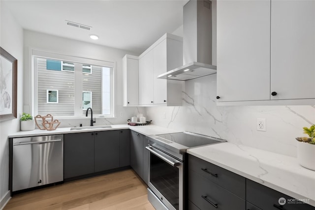 kitchen with light stone countertops, appliances with stainless steel finishes, sink, wall chimney range hood, and white cabinets