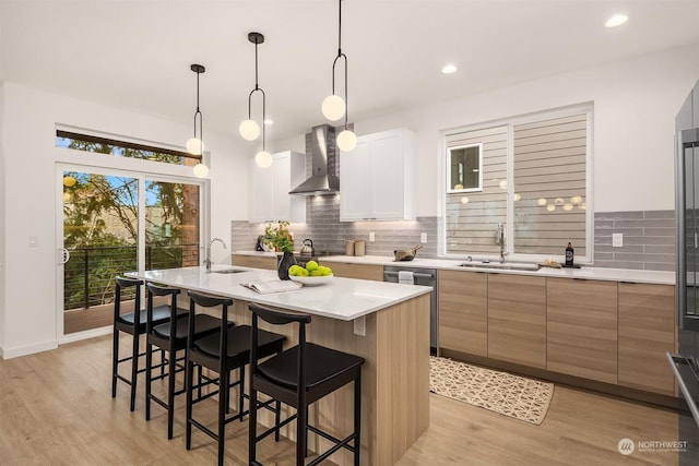 kitchen with stainless steel dishwasher, wall chimney range hood, pendant lighting, white cabinets, and an island with sink