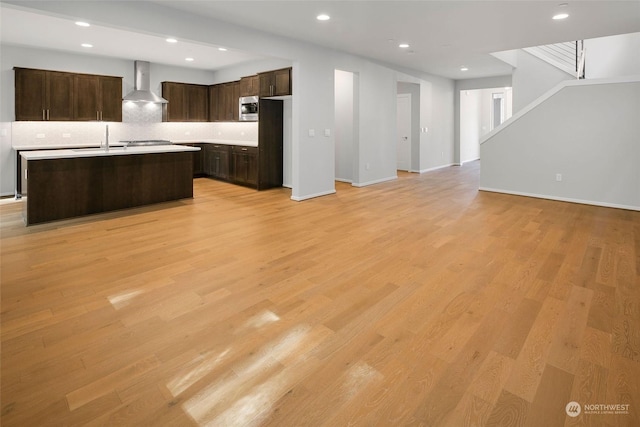 kitchen with stainless steel microwave, a center island with sink, wall chimney exhaust hood, dark brown cabinets, and light hardwood / wood-style floors