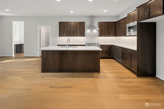 kitchen featuring sink, wall chimney range hood, a center island with sink, light hardwood / wood-style flooring, and stainless steel microwave