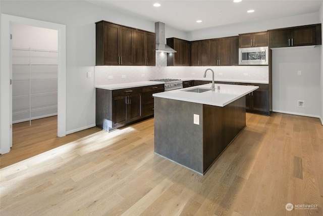 kitchen with stainless steel microwave, wall chimney exhaust hood, sink, a center island with sink, and light wood-type flooring