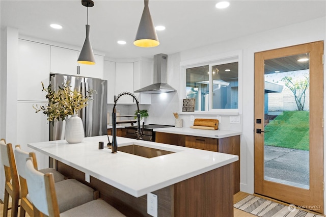 kitchen with pendant lighting, a center island with sink, wall chimney range hood, stainless steel fridge, and white cabinetry