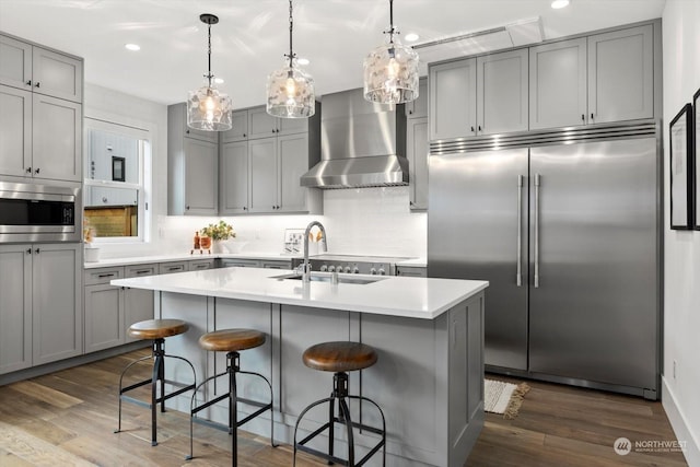 kitchen featuring a center island with sink, wall chimney range hood, hanging light fixtures, built in appliances, and dark hardwood / wood-style floors