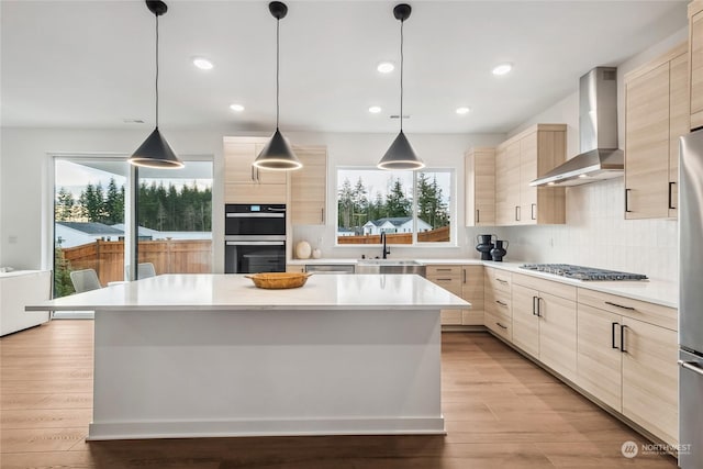 kitchen with appliances with stainless steel finishes, light brown cabinets, hanging light fixtures, and wall chimney exhaust hood