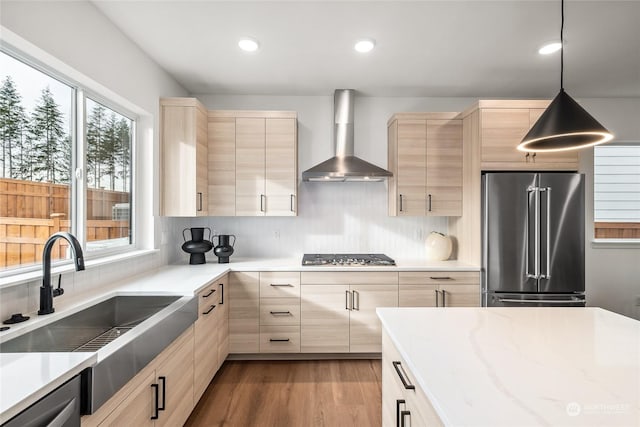 kitchen with sink, wall chimney exhaust hood, hanging light fixtures, stainless steel appliances, and light brown cabinetry