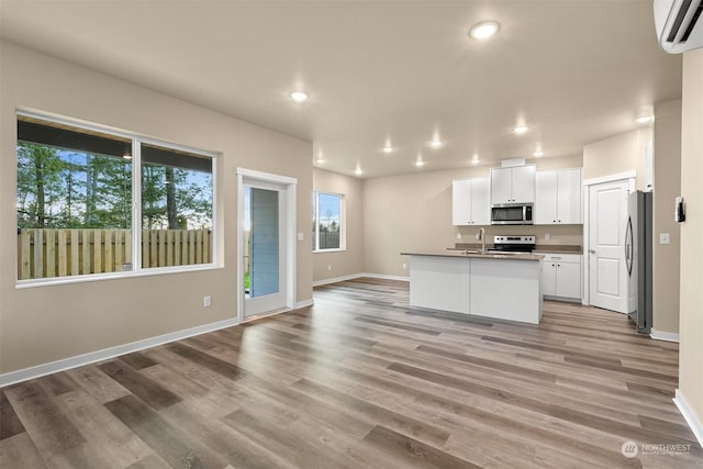 kitchen featuring a wall mounted air conditioner, a kitchen island with sink, appliances with stainless steel finishes, light hardwood / wood-style floors, and white cabinetry