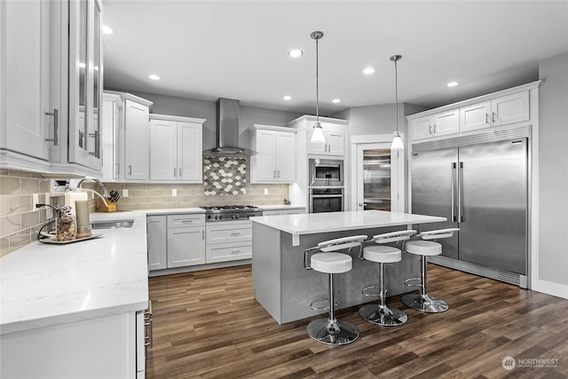 kitchen featuring a center island, white cabinets, hanging light fixtures, built in appliances, and wall chimney exhaust hood