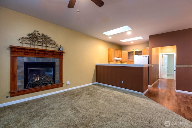 unfurnished living room with ceiling fan, carpet flooring, a skylight, and a tiled fireplace