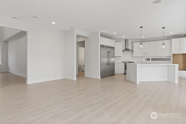 kitchen with wall chimney exhaust hood, hanging light fixtures, a kitchen island with sink, white cabinets, and appliances with stainless steel finishes