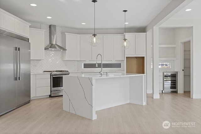 kitchen with wall chimney exhaust hood, wine cooler, a kitchen island with sink, white cabinets, and appliances with stainless steel finishes