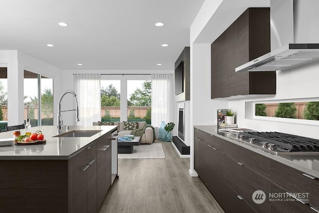 kitchen with sink, wall chimney range hood, stainless steel gas cooktop, light hardwood / wood-style floors, and a center island with sink