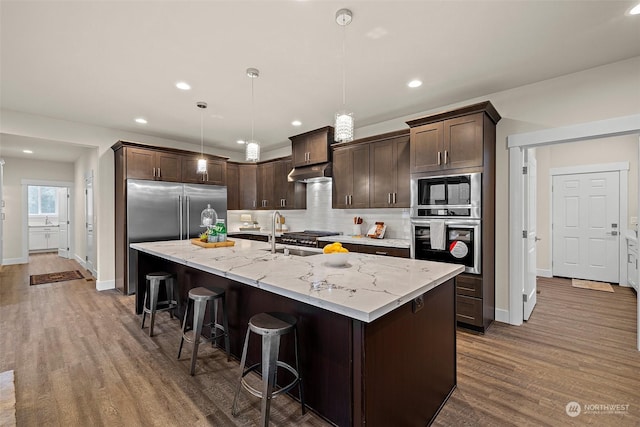 kitchen featuring decorative light fixtures, dark brown cabinetry, a kitchen island with sink, and built in appliances