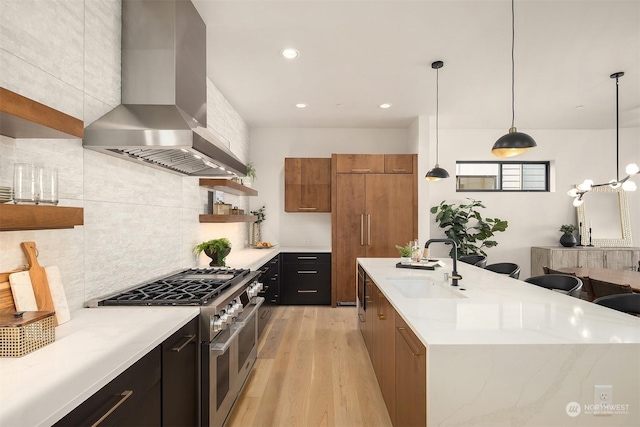 kitchen featuring sink, wall chimney range hood, range with two ovens, hanging light fixtures, and an island with sink