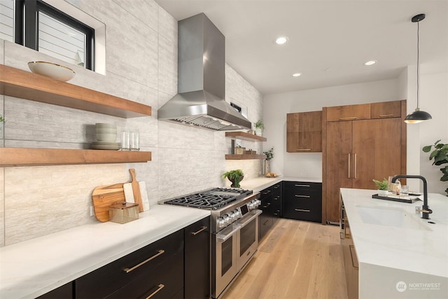 kitchen featuring double oven range, sink, wall chimney exhaust hood, decorative light fixtures, and light hardwood / wood-style floors