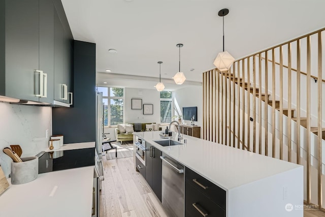 kitchen featuring light wood-type flooring, stainless steel dishwasher, a kitchen island with sink, sink, and hanging light fixtures