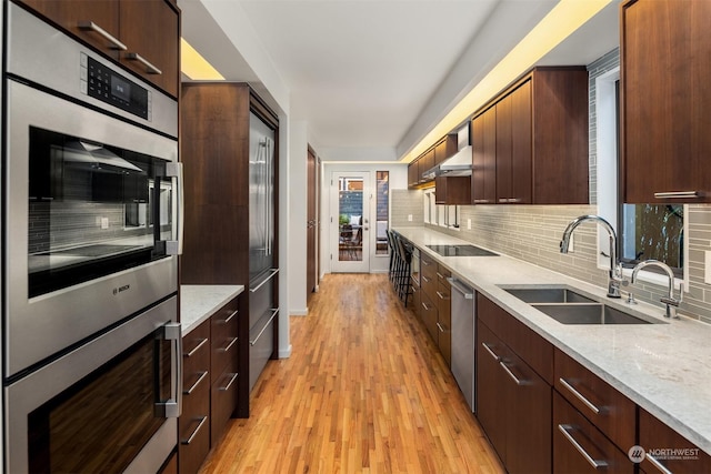 kitchen with appliances with stainless steel finishes, light wood-type flooring, light stone counters, sink, and tasteful backsplash