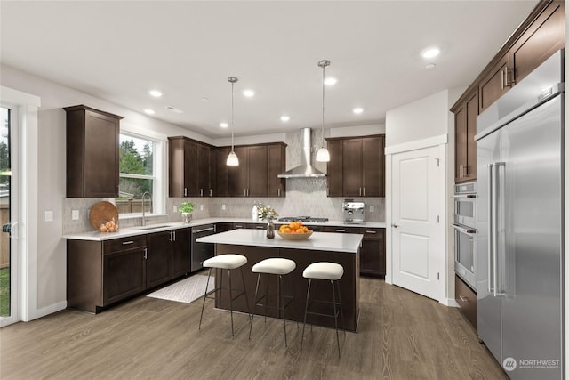 kitchen featuring stainless steel appliances, pendant lighting, a kitchen island, wall chimney exhaust hood, and dark brown cabinets