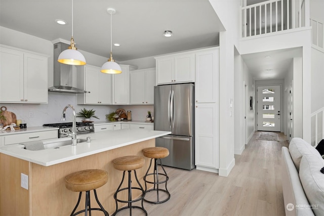 kitchen featuring stainless steel refrigerator, white cabinetry, hanging light fixtures, a kitchen island with sink, and wall chimney exhaust hood