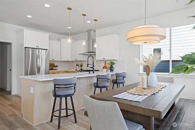 kitchen with hanging light fixtures, white cabinetry, wall chimney range hood, and stainless steel fridge