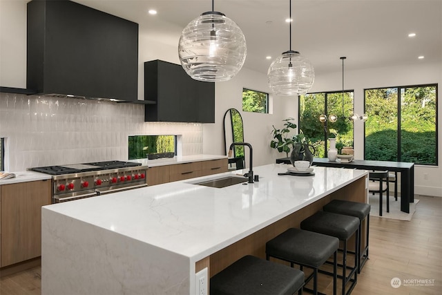 kitchen featuring backsplash, a kitchen island with sink, decorative light fixtures, range with two ovens, and range hood