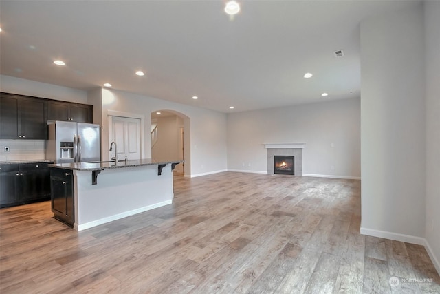 kitchen featuring arched walkways, stainless steel fridge with ice dispenser, light wood-style flooring, a breakfast bar area, and dark stone countertops