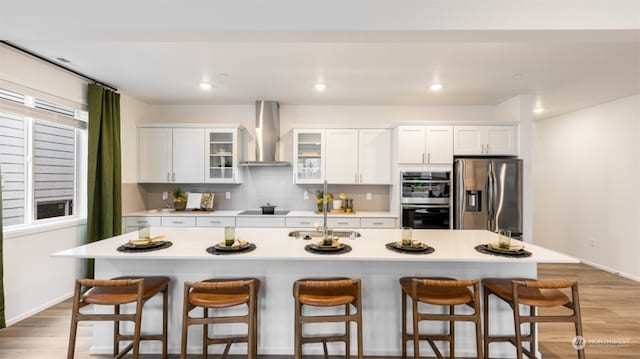 kitchen with white cabinets, wall chimney range hood, stainless steel appliances, and a kitchen island with sink