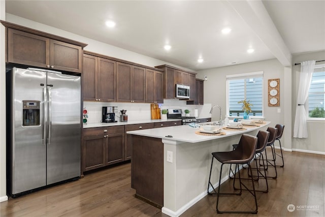 kitchen featuring appliances with stainless steel finishes, tasteful backsplash, dark brown cabinetry, a center island with sink, and dark wood-type flooring