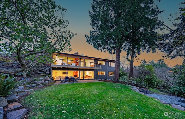 back house at dusk with a balcony, a patio area, and a yard