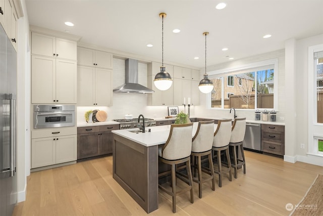 kitchen featuring wall chimney exhaust hood, a kitchen breakfast bar, light hardwood / wood-style floors, a kitchen island with sink, and appliances with stainless steel finishes