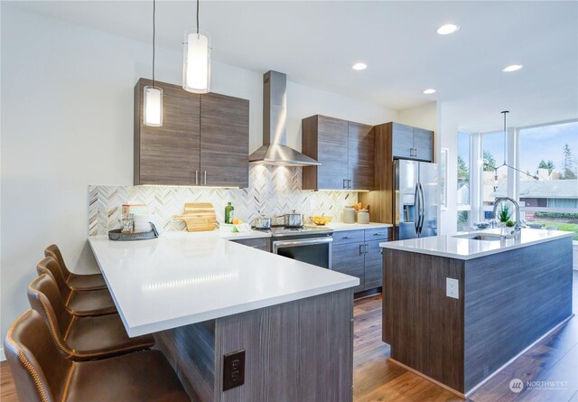 kitchen with a kitchen bar, appliances with stainless steel finishes, decorative backsplash, hanging light fixtures, and wall chimney range hood