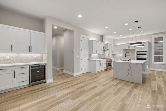 kitchen featuring wall chimney exhaust hood, hanging light fixtures, a kitchen island with sink, wine cooler, and appliances with stainless steel finishes